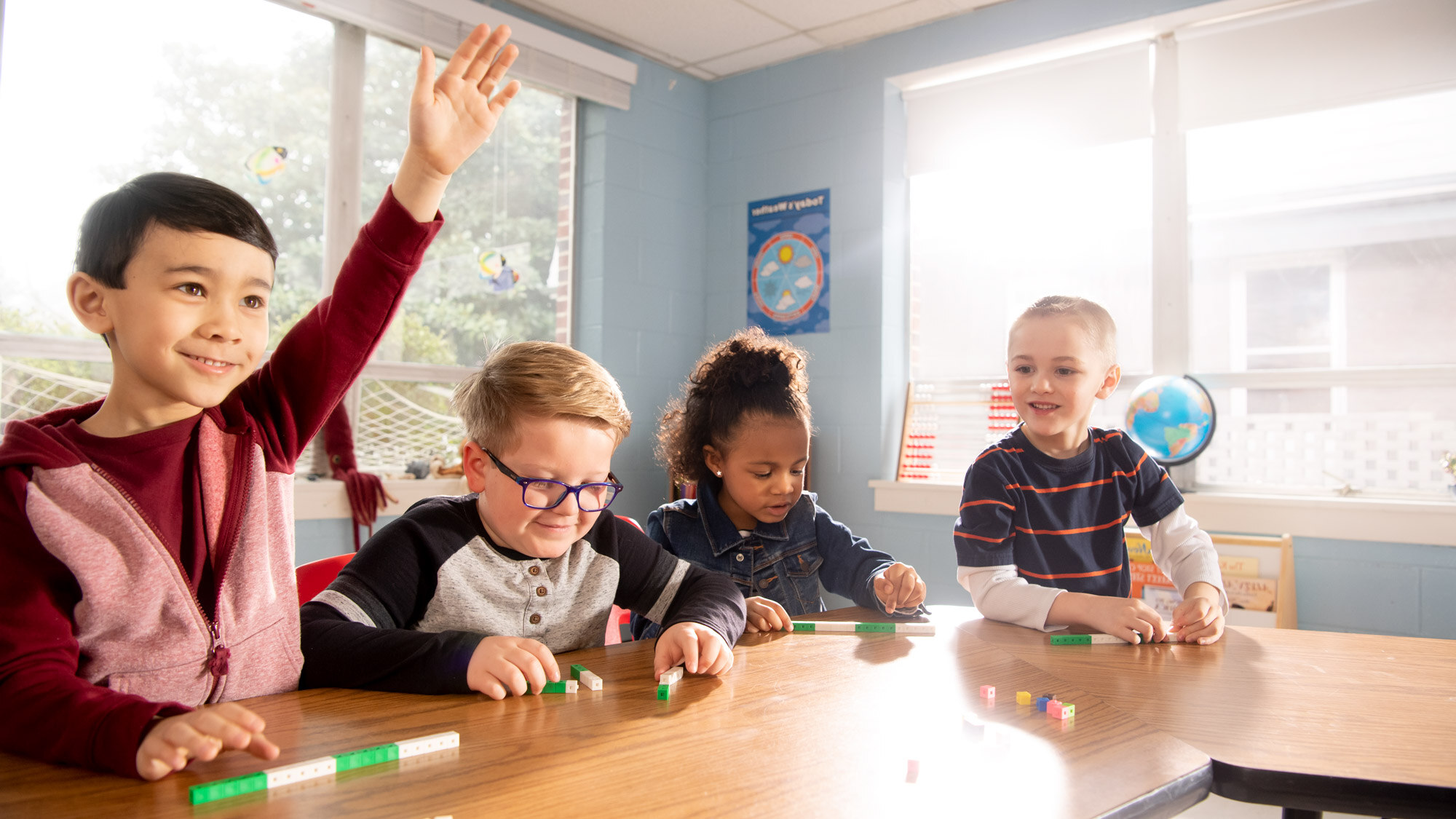 Four students sit around a table with block in front of them. One student is raising their hand.