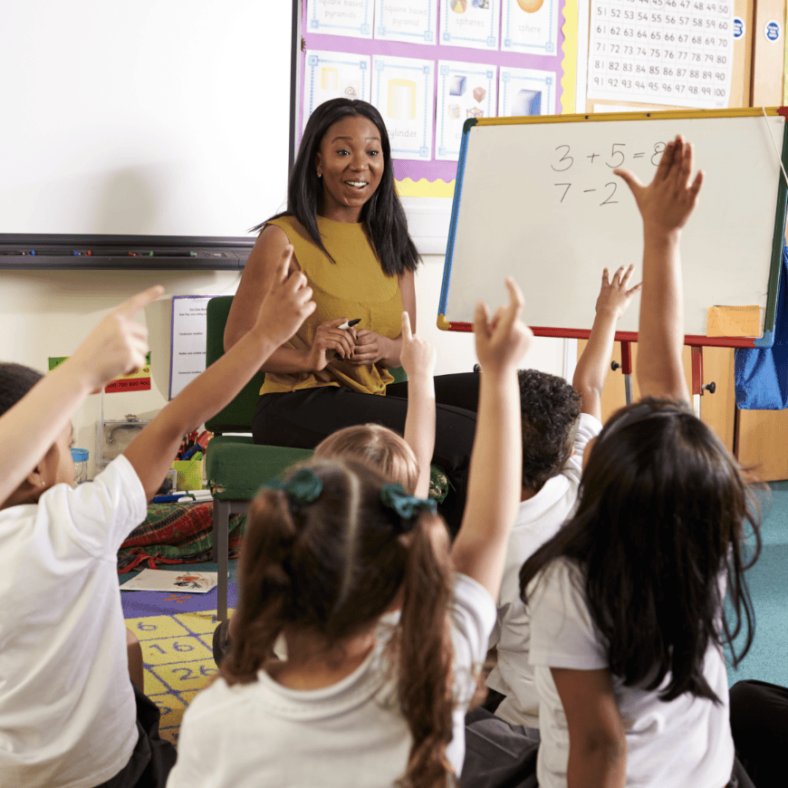 Students sitting on the carpet in front of their teacher. Students raise hands as the teachers writes number sentences on the whiteboard.